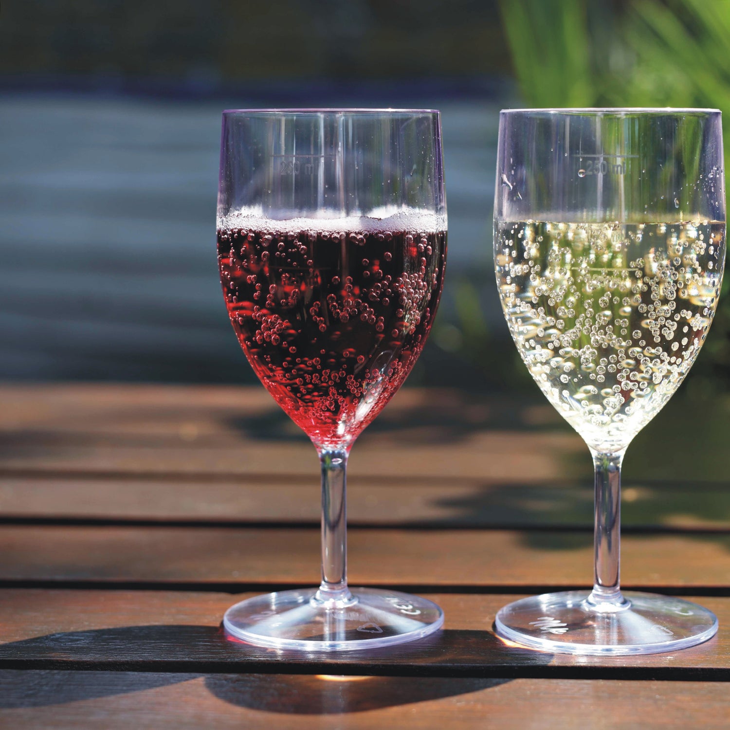 Two plastic wine glasses each filled with a red and light coloured beverage sitting on a wooden table in an outdoor garden setting