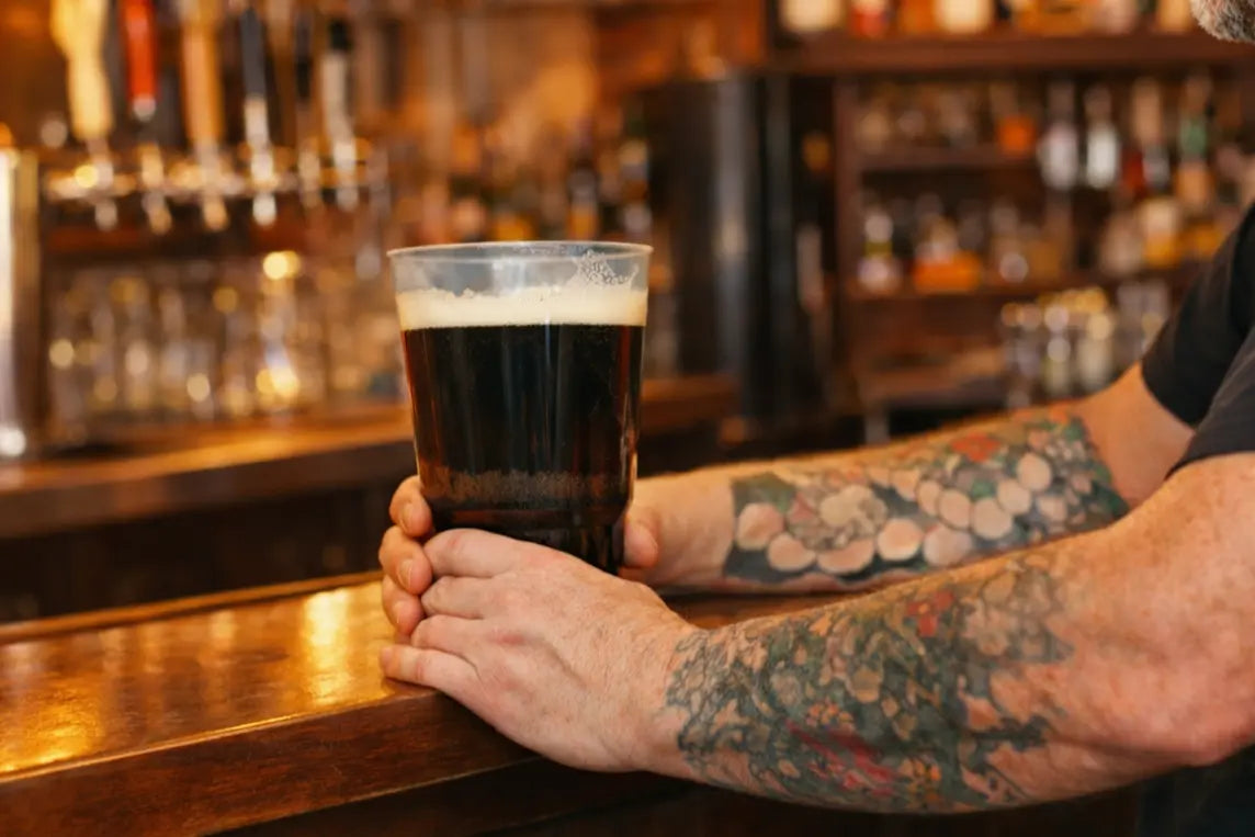 Person holding a glass of dark beer in a plastic pint cup bar setting