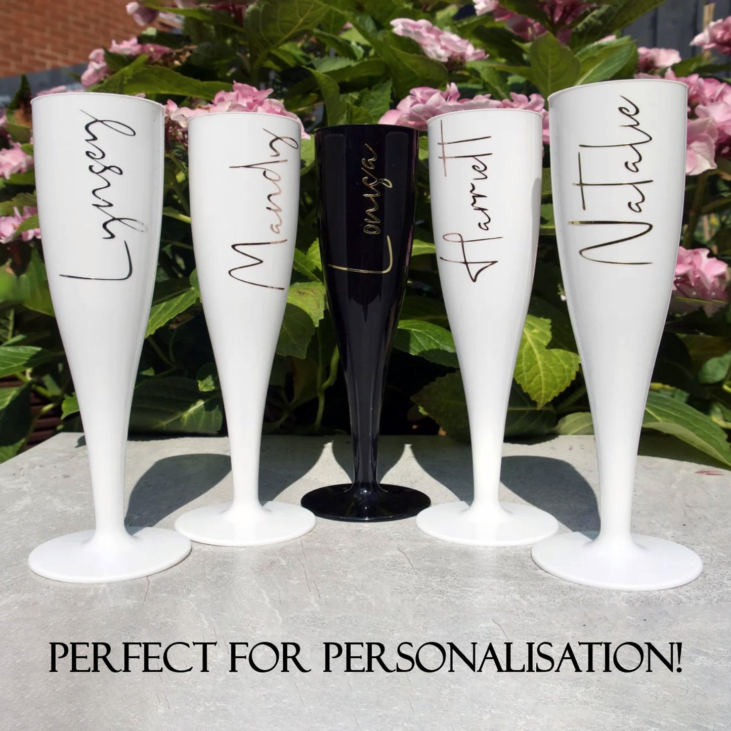 One black and four white champagne flutes personalised with names in a vinyl graphic with a garden bush in the background