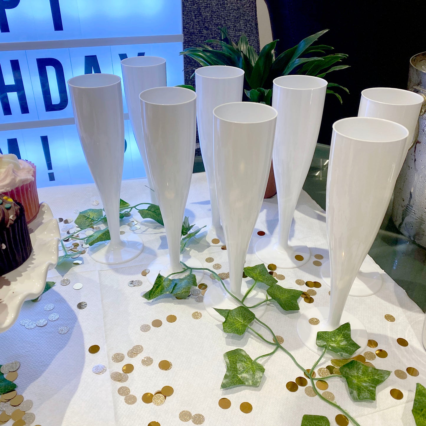 Several white champagne flutes on a table decorated with gold and silver confetti with a light board in the background and cakes to the side